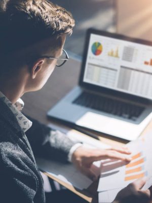 Young finance market analyst in eyeglasses working at sunny office on laptop while sitting at wooden table.Businessman analyze document in his hands.Graphs and diagramm on notebook screen.Vertical